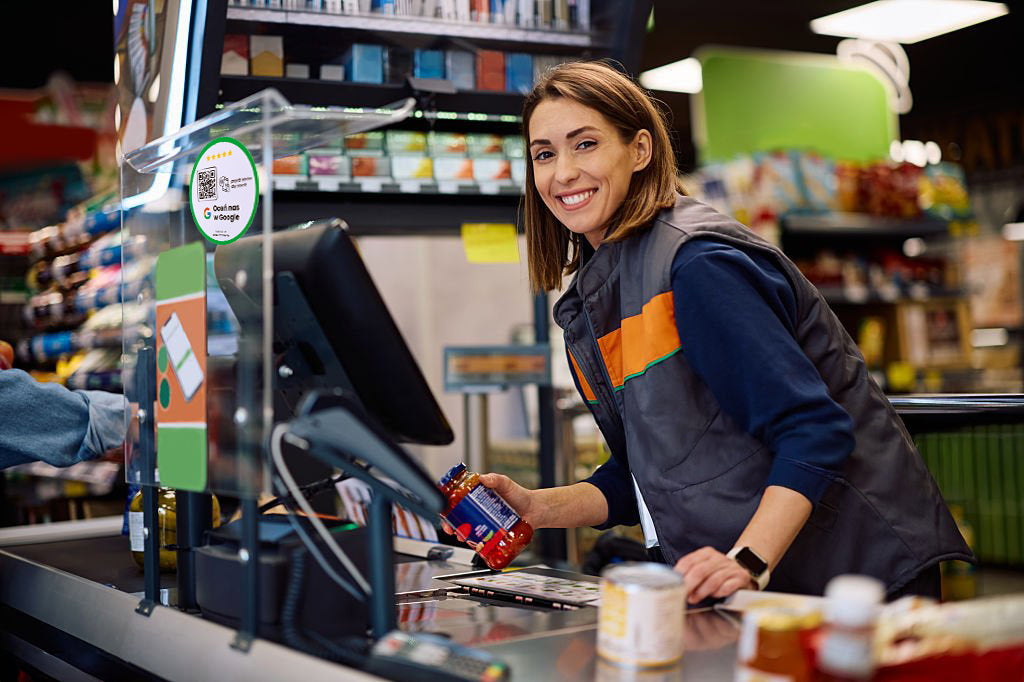 Uśmiechnięta kasjerka przy kasie z naklejką NFC/QR zachęcającą do zostawienia opinii